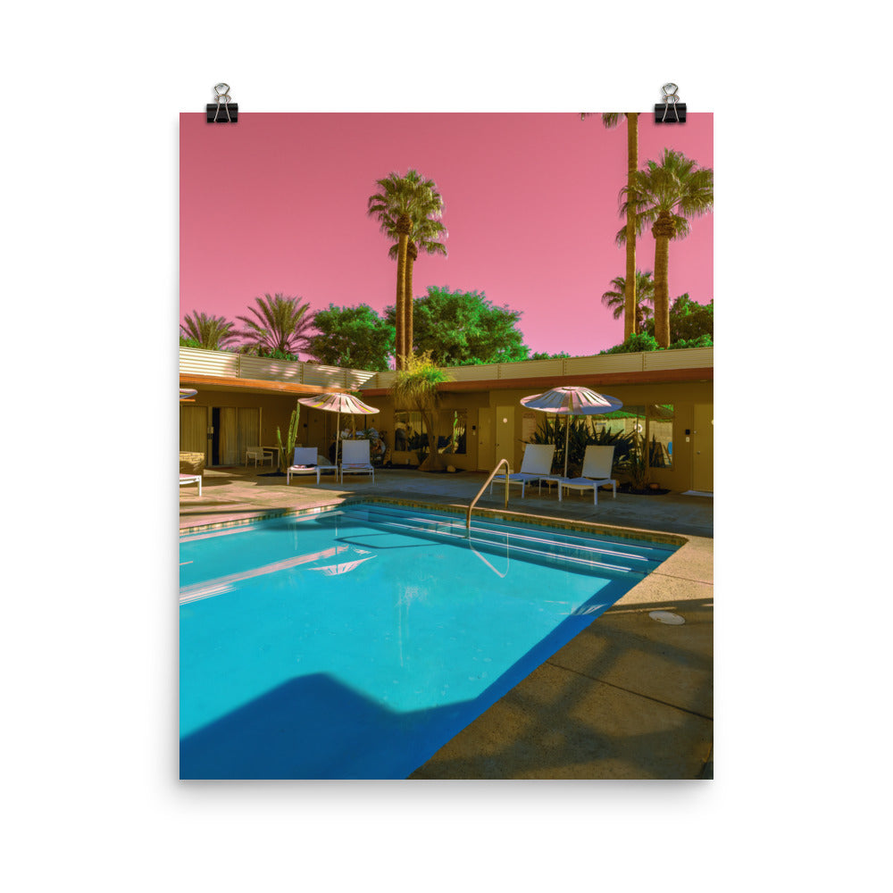 Framed fine art print of a retro Palm Springs motel pool, candy-pink sky above mid-century architecture, turquoise water reflecting striped pool umbrellas and white loungers, surrounded by tall palms and desert greenery.